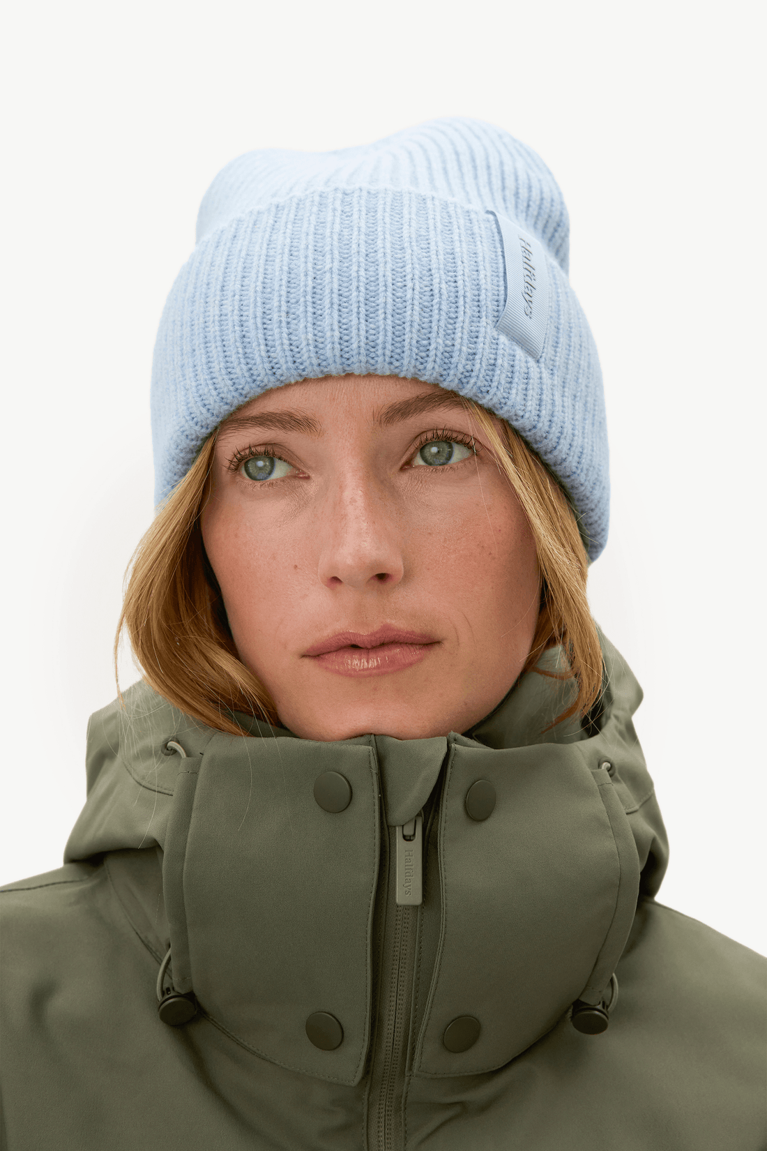 A close-up portrait of a woman wearing a light blue ribbed knit beanie and an olive green high-collar winter jacket against a plain white background.