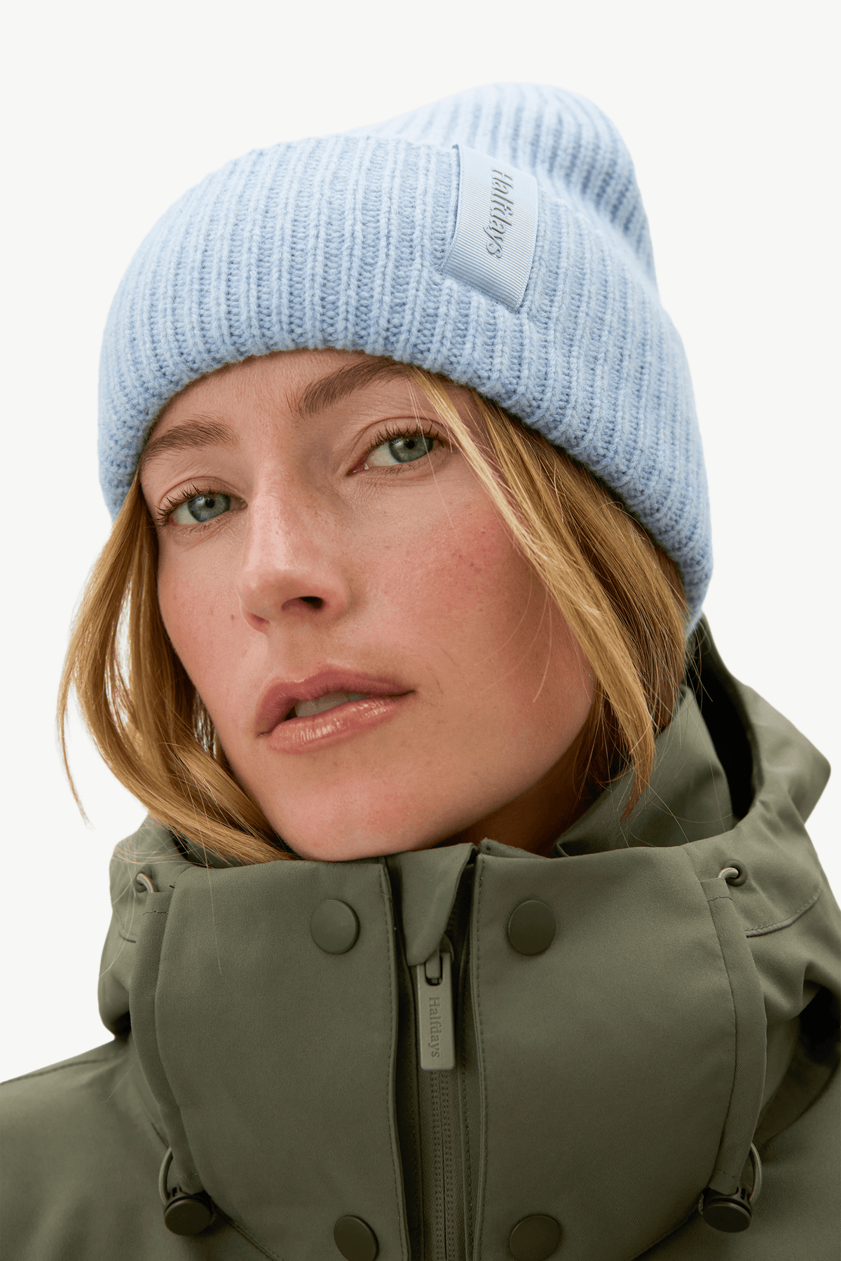 A close-up portrait of a woman wearing a light blue ribbed knit beanie and an olive green high-collar winter jacket against a plain white background.