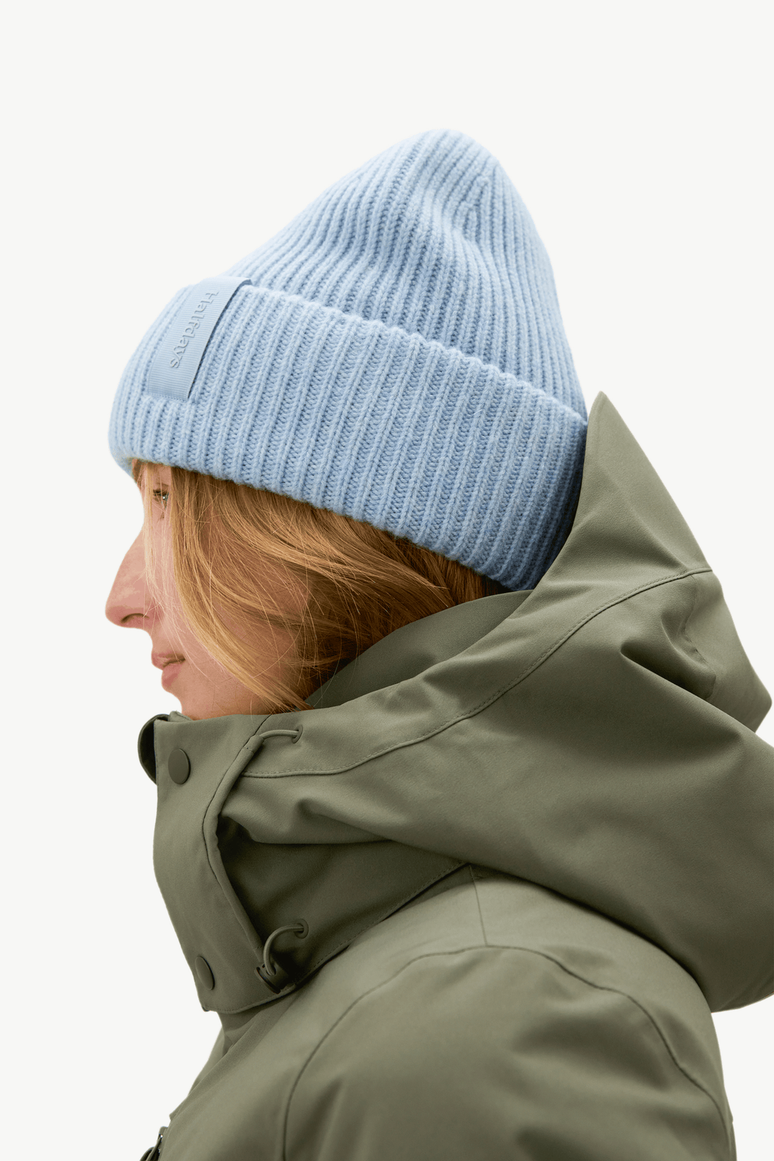 A close-up portrait of a woman wearing a light blue ribbed knit beanie and an olive green high-collar winter jacket against a plain white background.