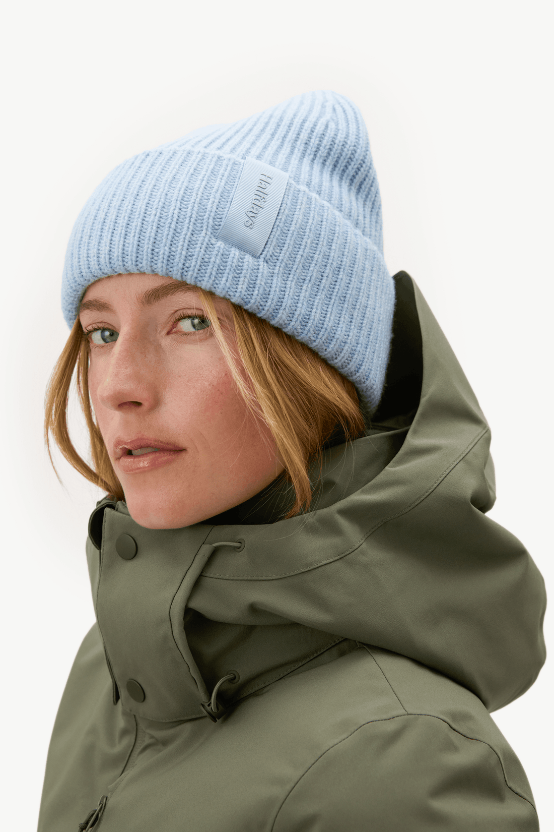 A close-up portrait of a woman wearing a light blue ribbed knit beanie and an olive green high-collar winter jacket against a plain white background.