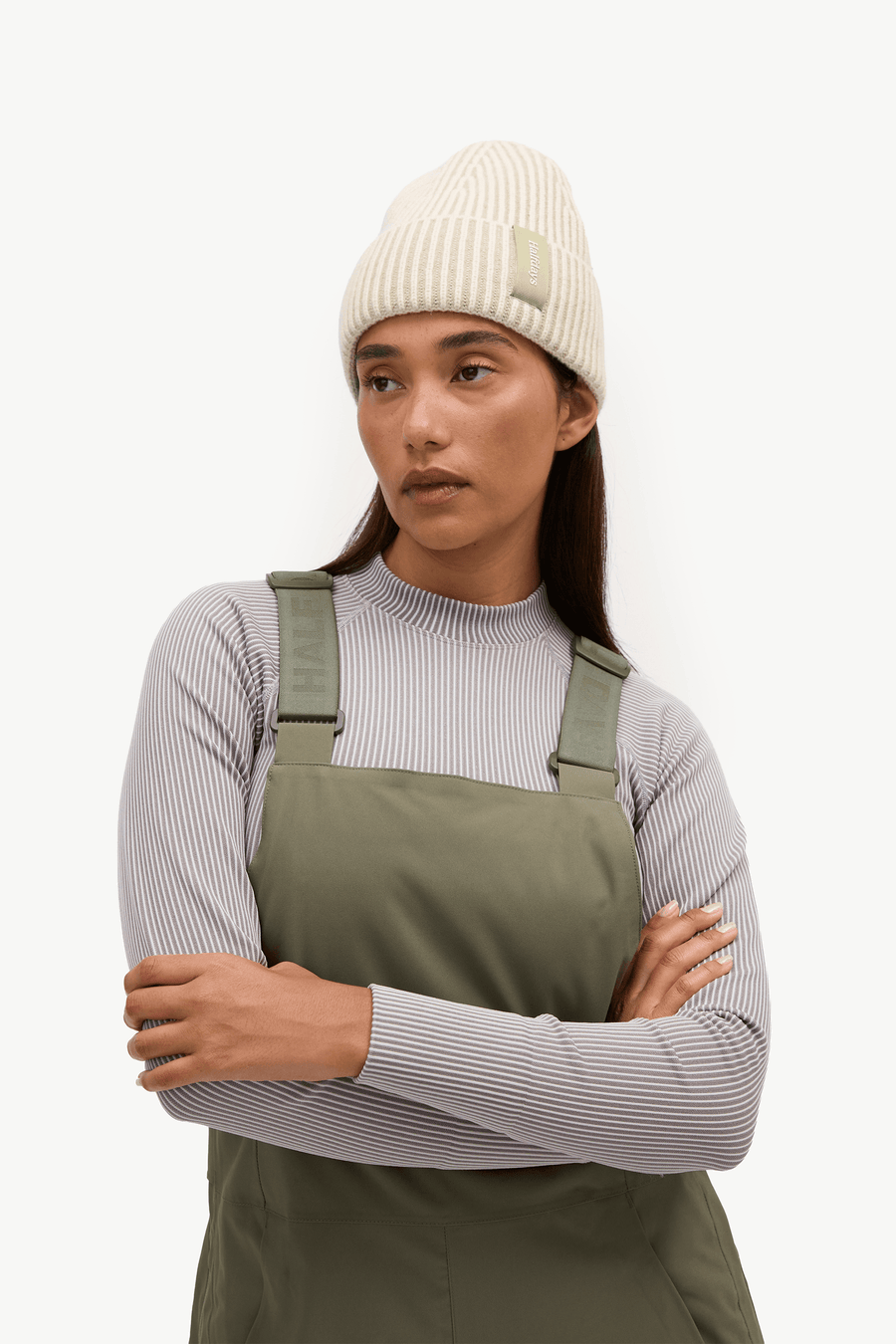 A woman wearing an olive green bib overalls over a light gray ribbed long-sleeve top, paired with a cream ribbed knit beanie, posed against a solid off-white background.