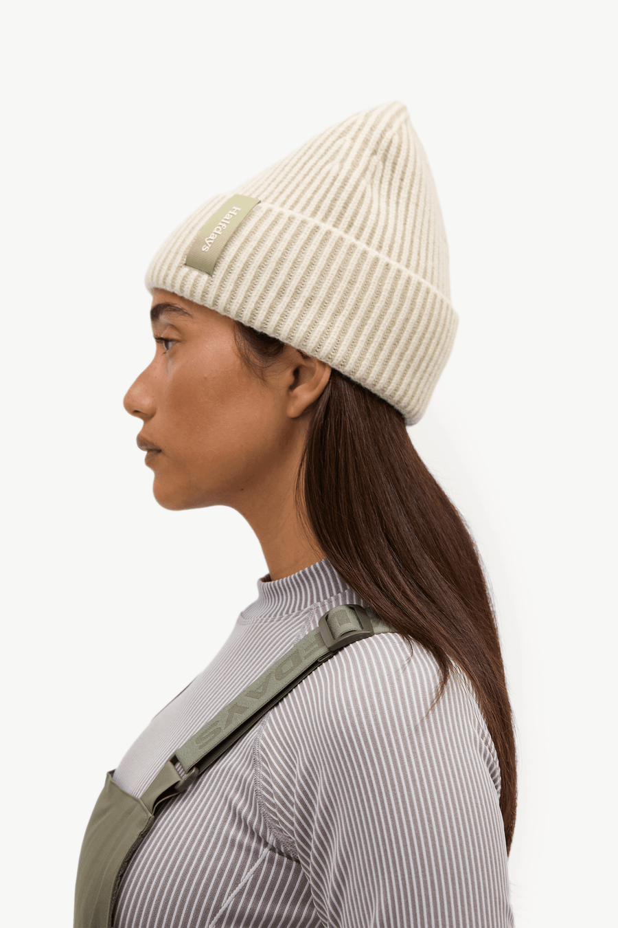 A woman wearing an olive green bib overalls over a light gray ribbed long-sleeve top, paired with a cream ribbed knit beanie, posed against a solid off-white background.