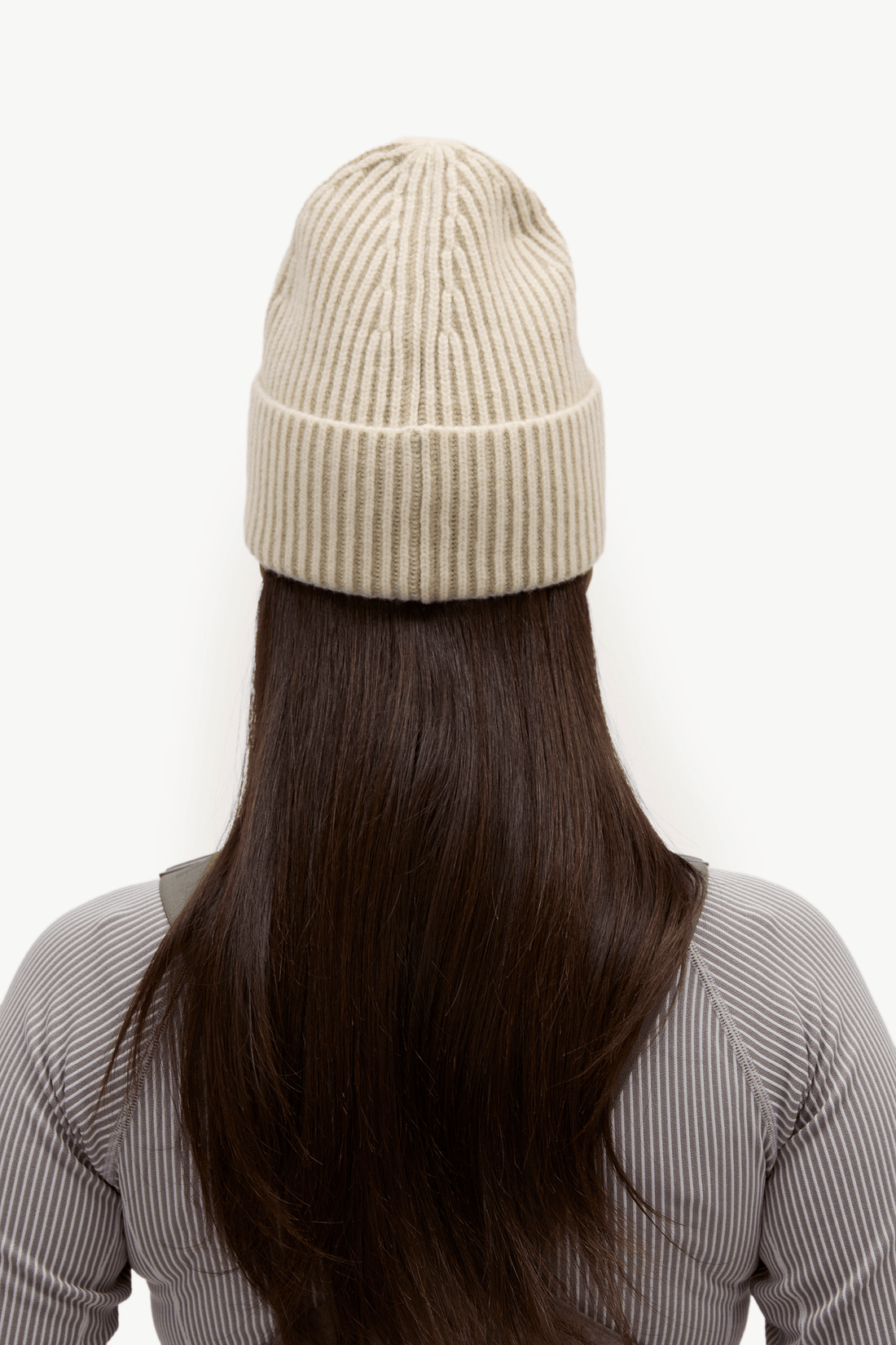 A woman wearing an olive green bib overalls over a light gray ribbed long-sleeve top, paired with a cream ribbed knit beanie, posed against a solid off-white background.