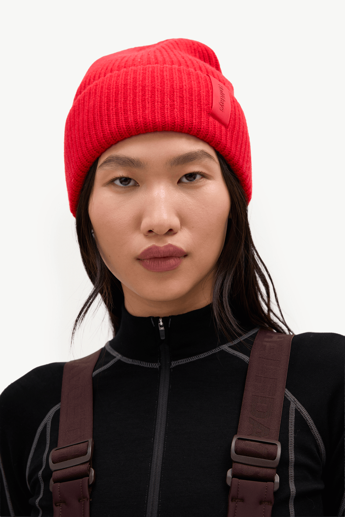 A close-up of a model wearing a vibrant red ribbed knit beanie hat and a black athletic zip-up top with brown harness straps, set against a plain light gray background.