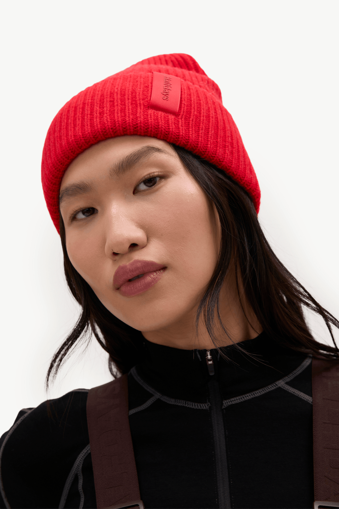 A close-up of a model wearing a bright red ribbed knit beanie with a small rectangular brand patch on the side, paired with a black top and brown shoulder straps against a plain white background.