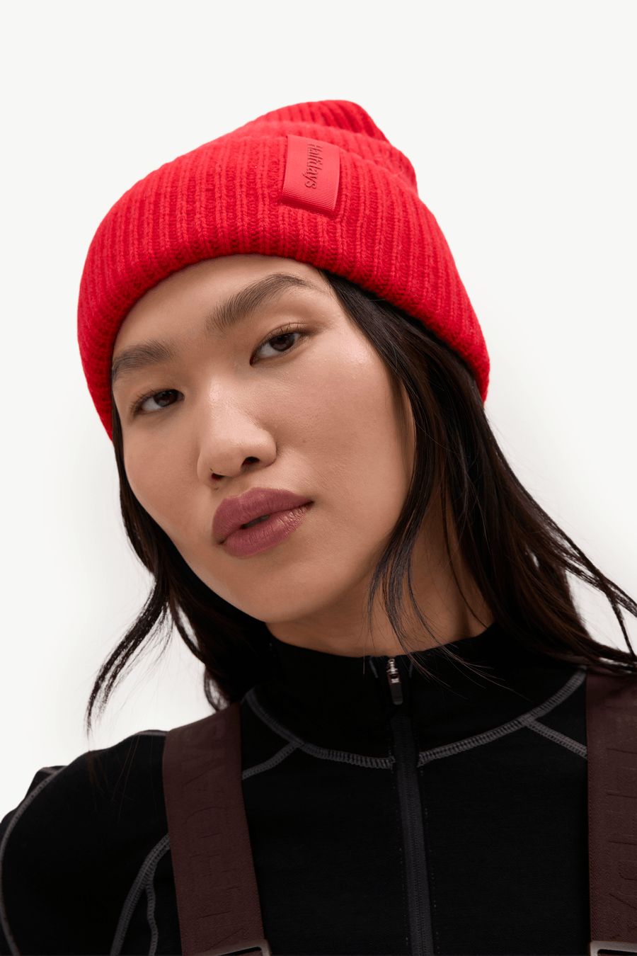 A close-up of a model wearing a bright red ribbed knit beanie with a small rectangular brand patch on the side, paired with a black top and brown shoulder straps against a plain white background.