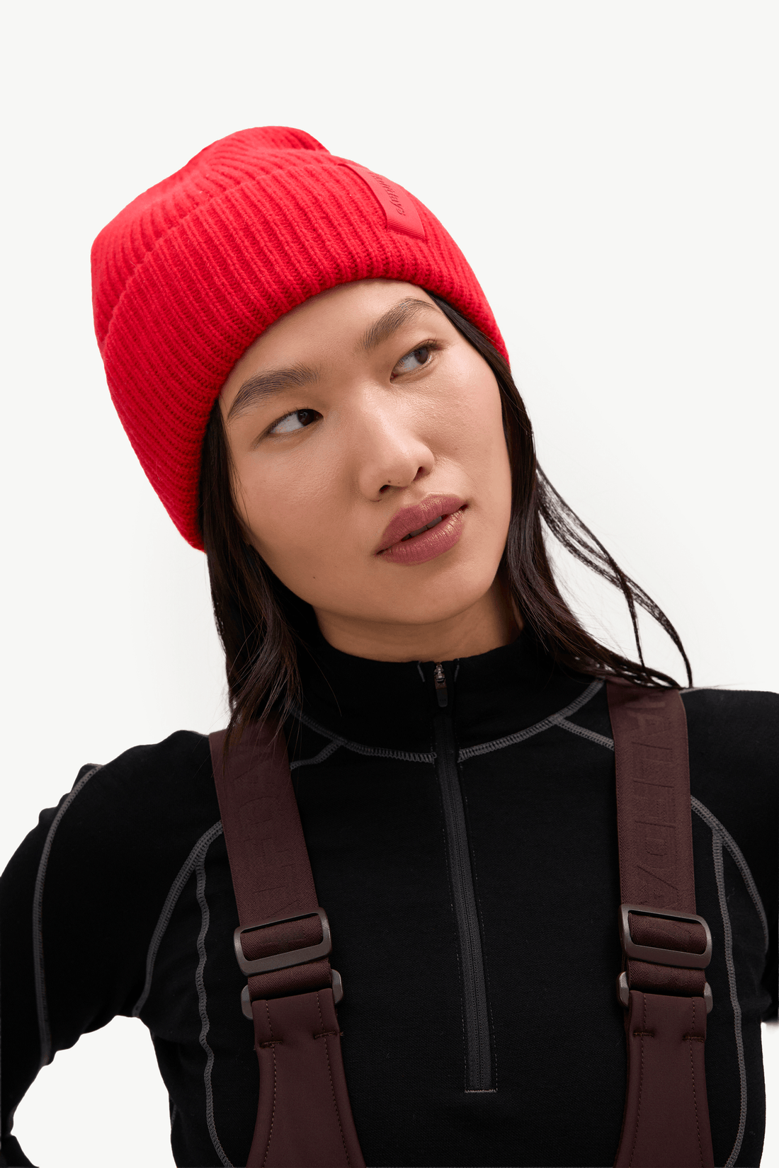 A woman wearing a bright red ribbed knit beanie and a black zip-up top with brown suspenders, photographed against a clean white studio background.