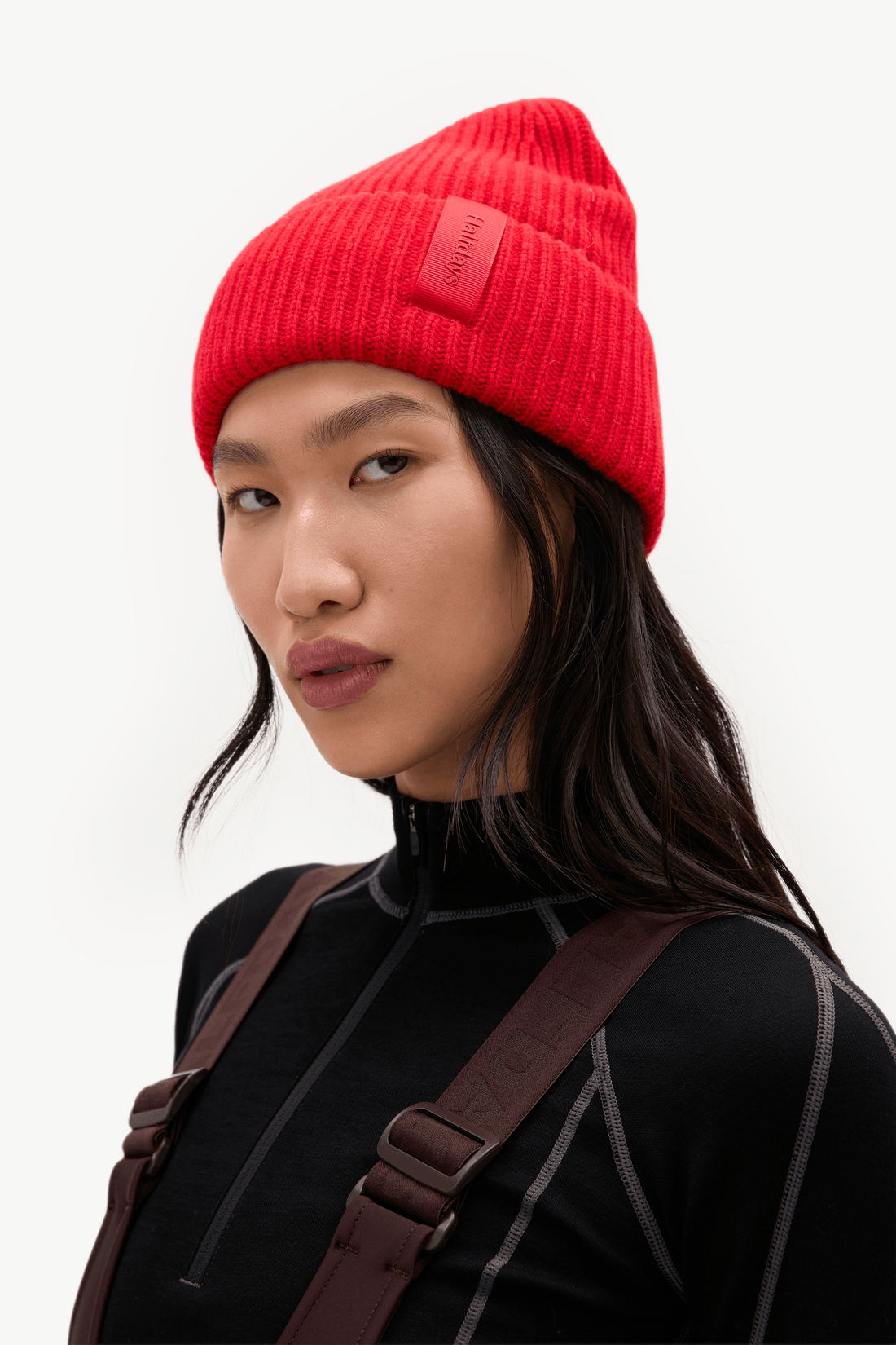 A model wearing a bright red ribbed knit beanie with a small rectangular brand patch on the side, paired with a black top and brown overalls against a clean white background.