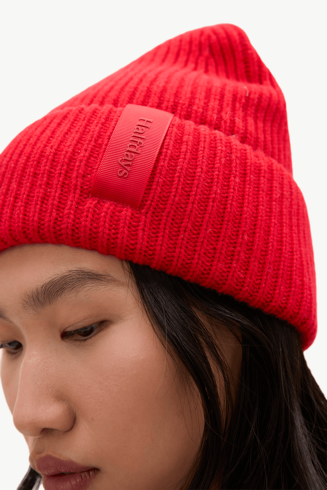 A close-up of a person wearing a bright red ribbed knit beanie with a matching red leather patch on the side featuring the debossed brand name 'HAIRBYES'.