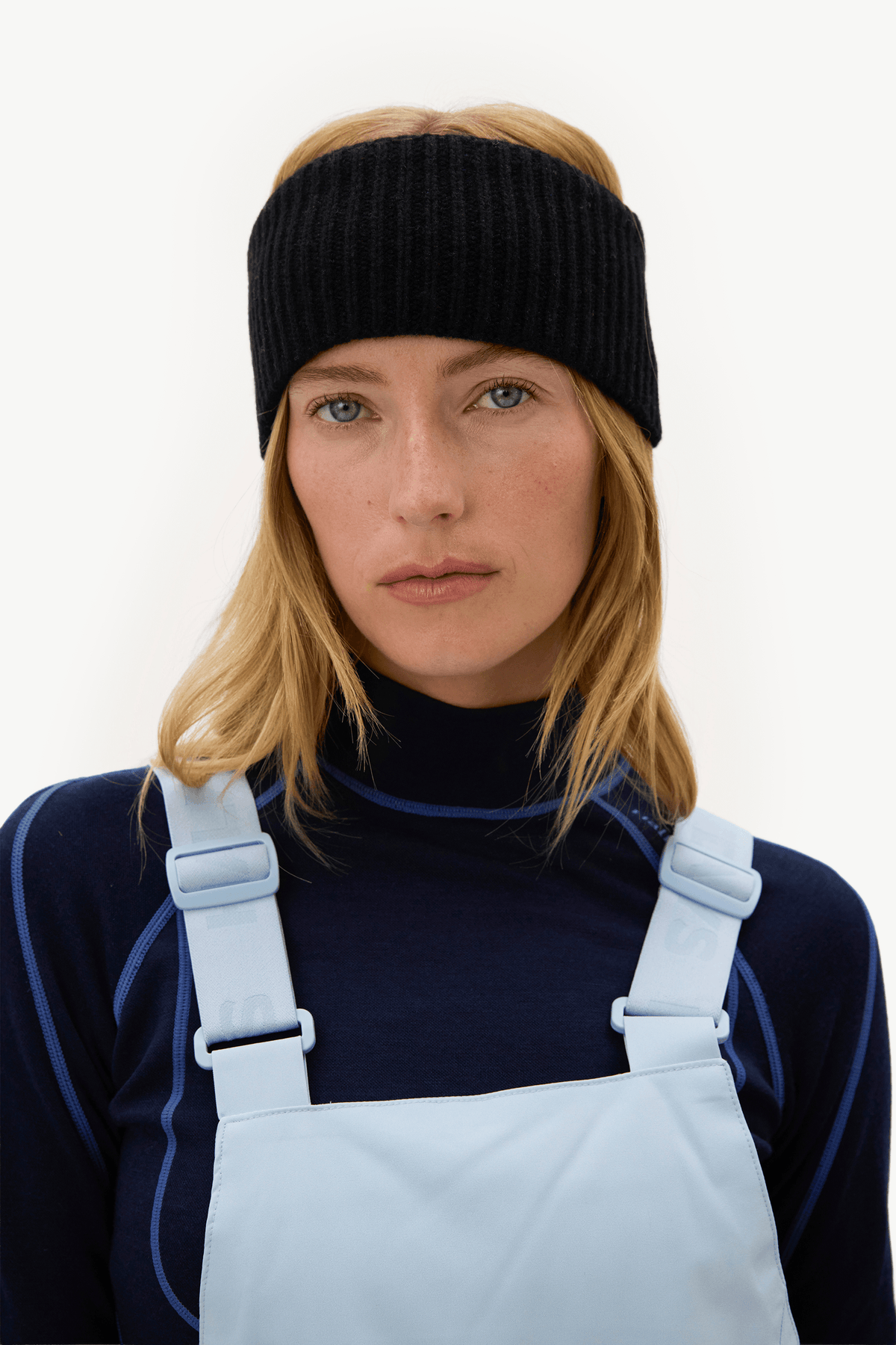 A woman wearing a black ribbed knit headband, a navy long-sleeve top, and light blue overalls against a plain white background.