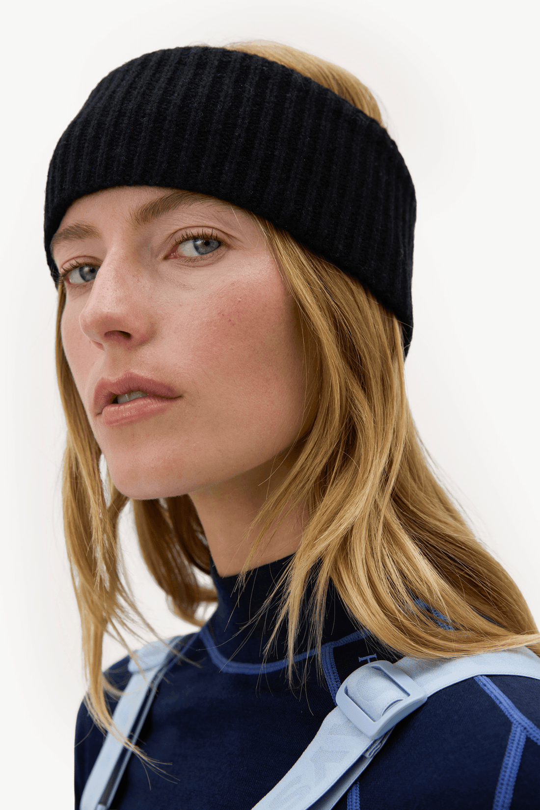 A woman wearing a black ribbed knit headband, a navy long-sleeve top, and light blue overalls against a plain white background.