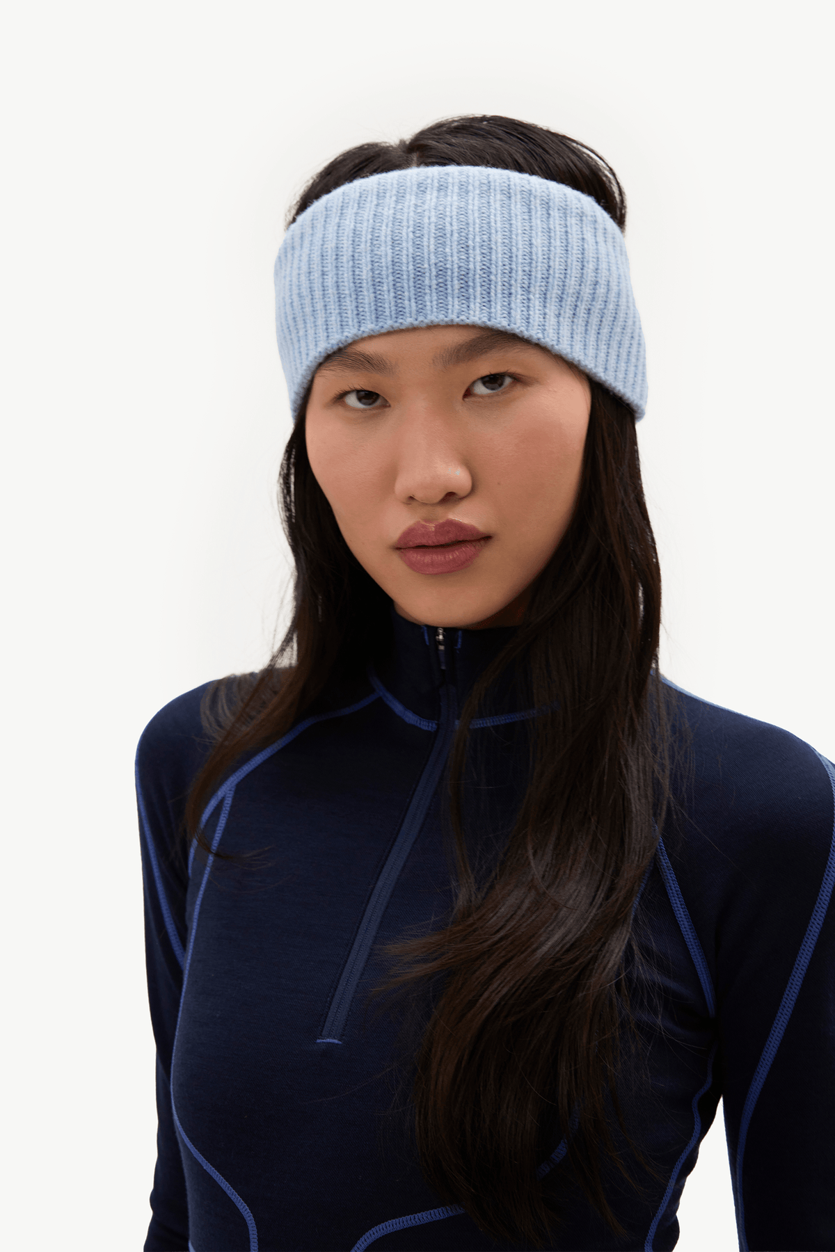 A woman wearing a light blue ribbed knit headband and a dark navy athletic quarter-zip top, photographed against a clean white studio background.