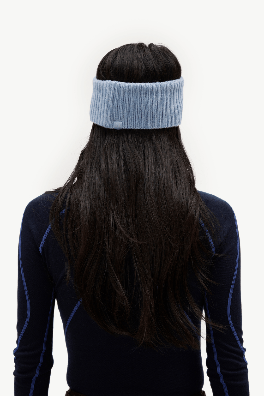 A person viewed from the back wearing a light blue ribbed knit headband and a dark navy long-sleeve athletic top with blue piping, against a plain white background.