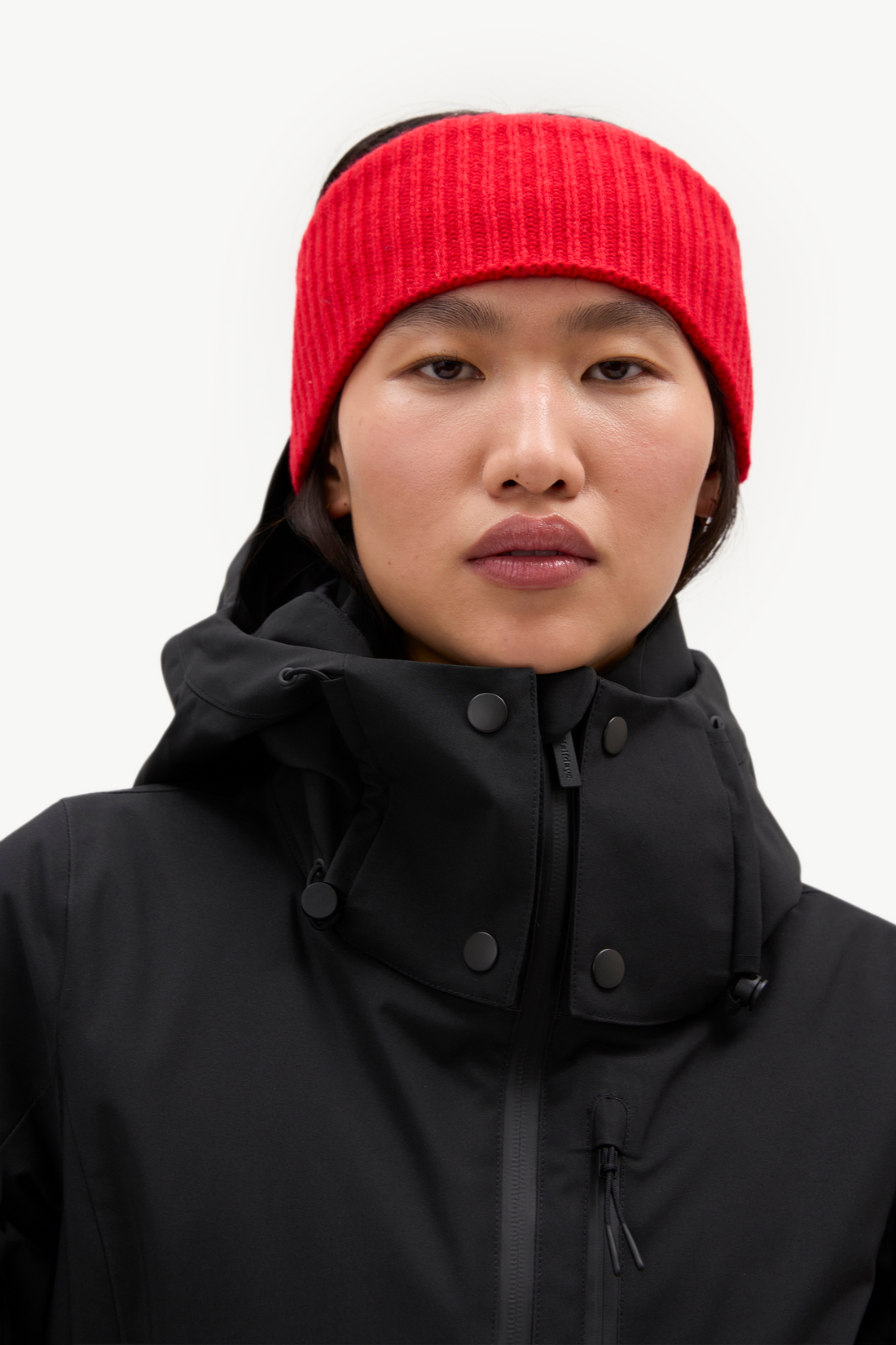 A close-up of a model wearing a bright red ribbed knit headband and a black high-collar winter jacket against a clean white background.