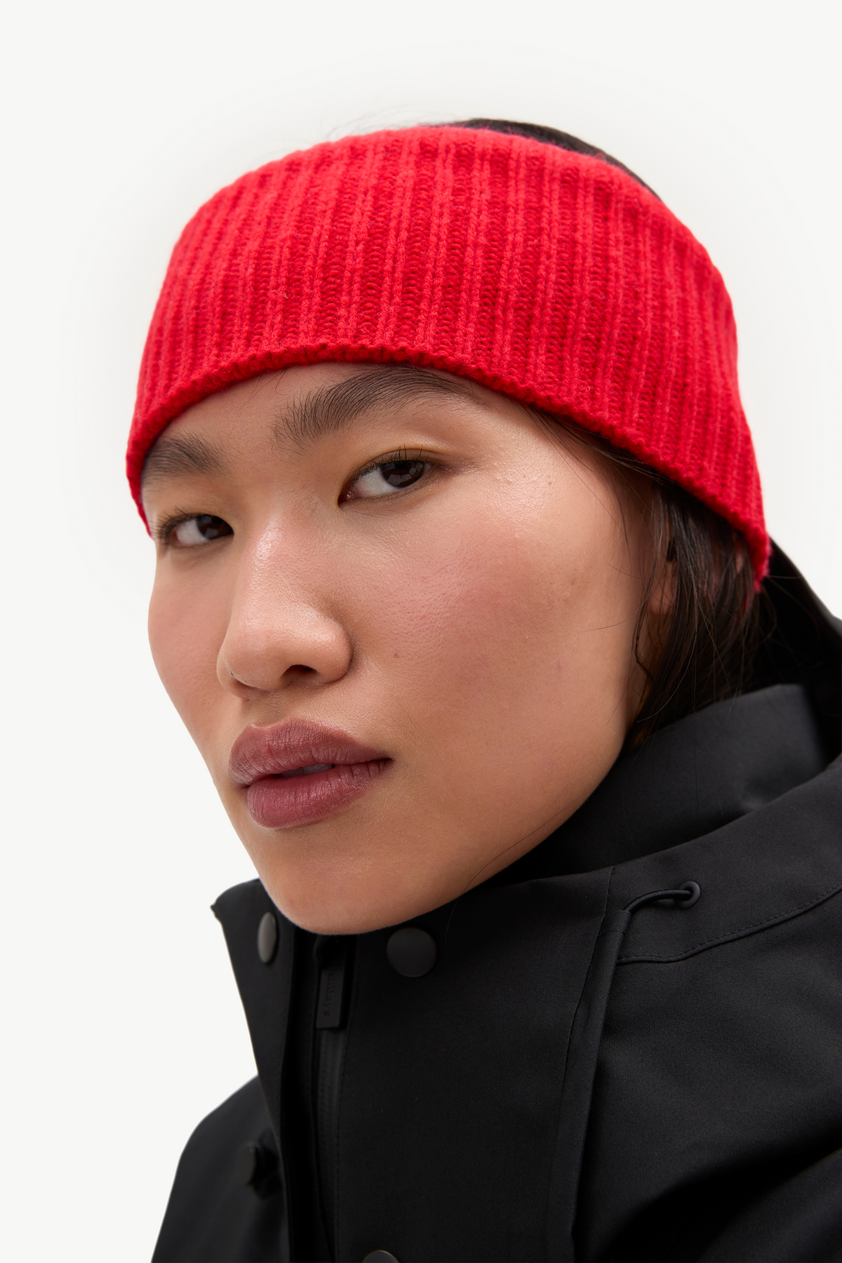 A close-up portrait of a model wearing a bright red ribbed knit headband and a black jacket against a clean white background.