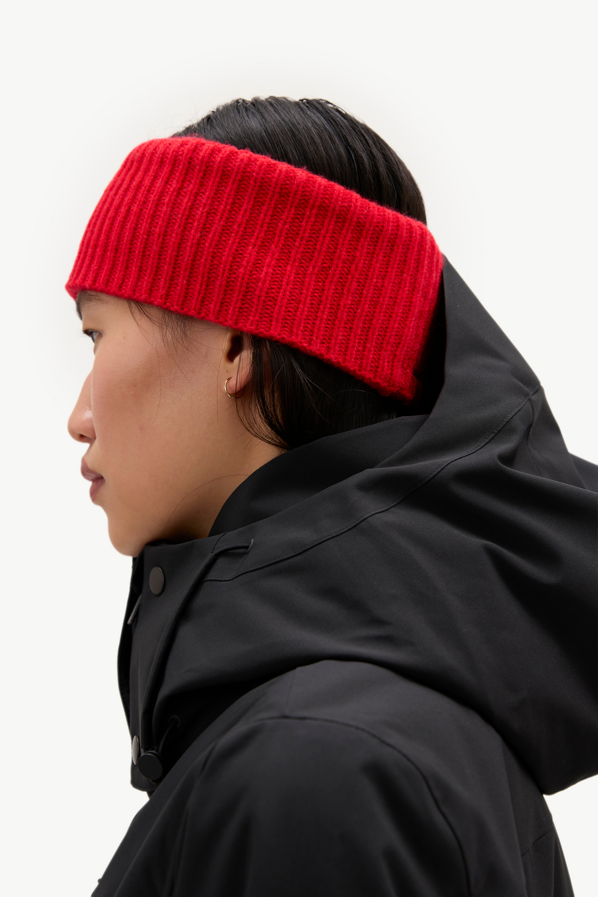 A side-profile view of a person wearing a bright red ribbed knit headband and a black hooded jacket against a clean white background.