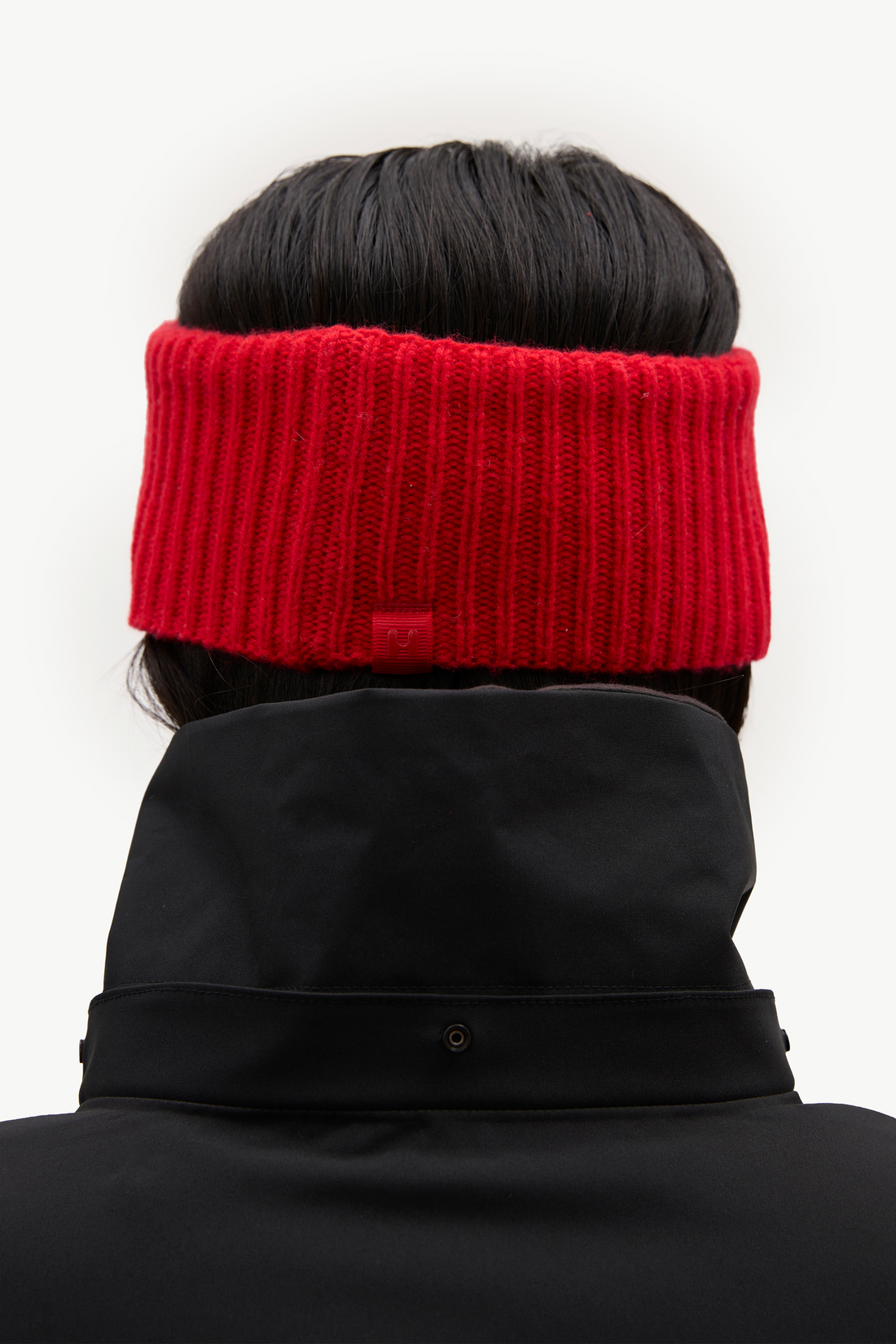 A close-up back view of a person wearing a bright red ribbed knit headband and a black high-collar jacket against a plain white background.