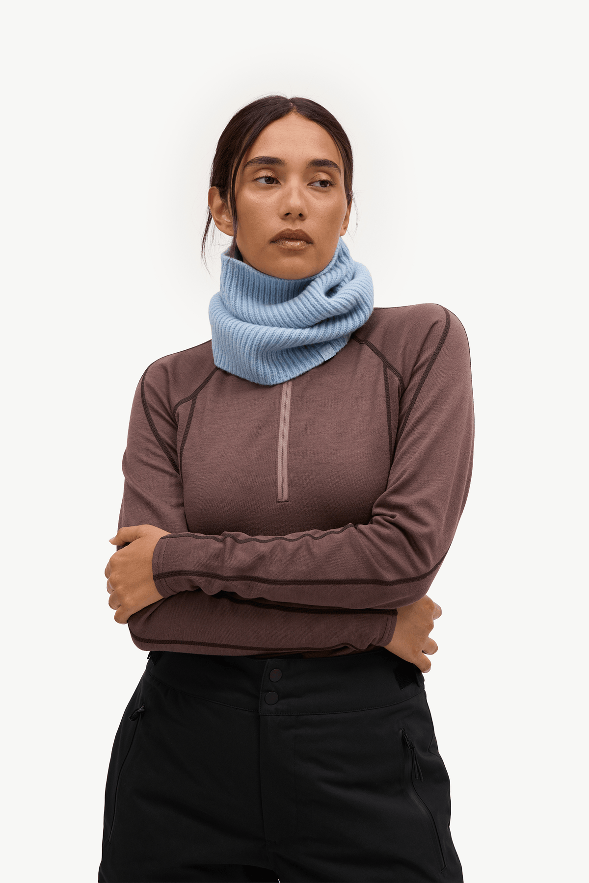 A woman wearing a light blue ribbed knit neck warmer cowl over a brown long-sleeve quarter-zip athletic top and black pants, photographed against a clean white studio background.