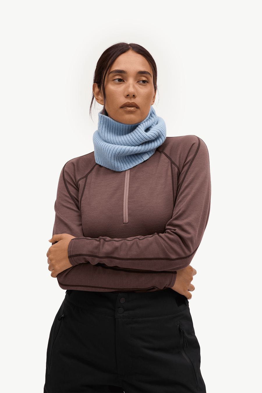 A woman wearing a light blue ribbed knit neck warmer cowl over a brown long-sleeve quarter-zip athletic top and black pants, photographed against a clean white studio background.