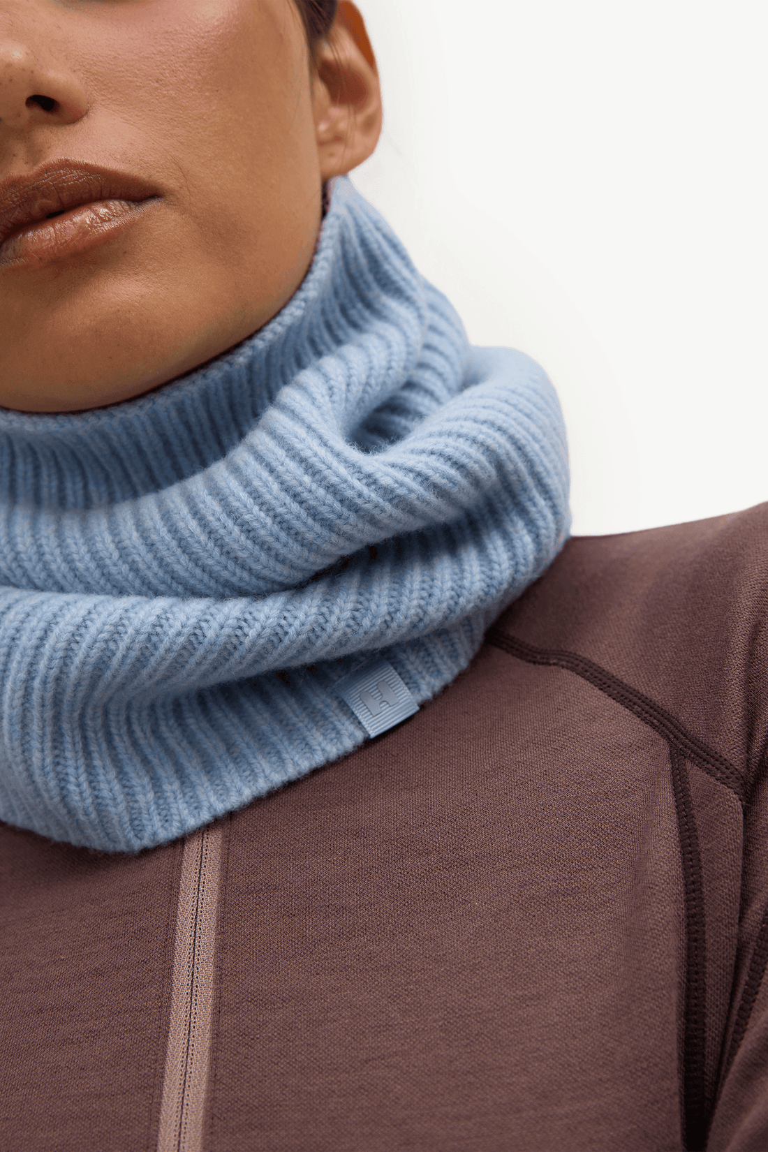 A close-up of a person wearing a light blue ribbed knit cowl neck warmer over a brown athletic top, against a plain white background.