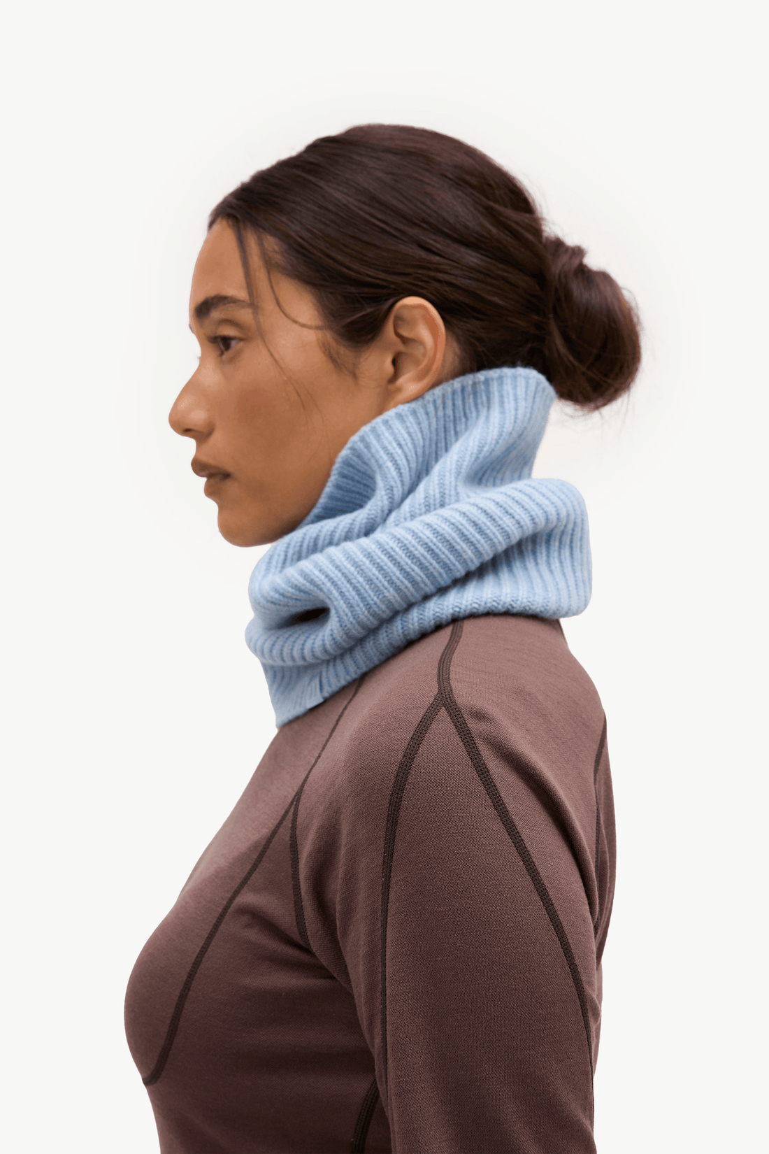 A woman in profile wearing a light blue ribbed knit cowl neck warmer over a brown athletic top, photographed against a clean white background.