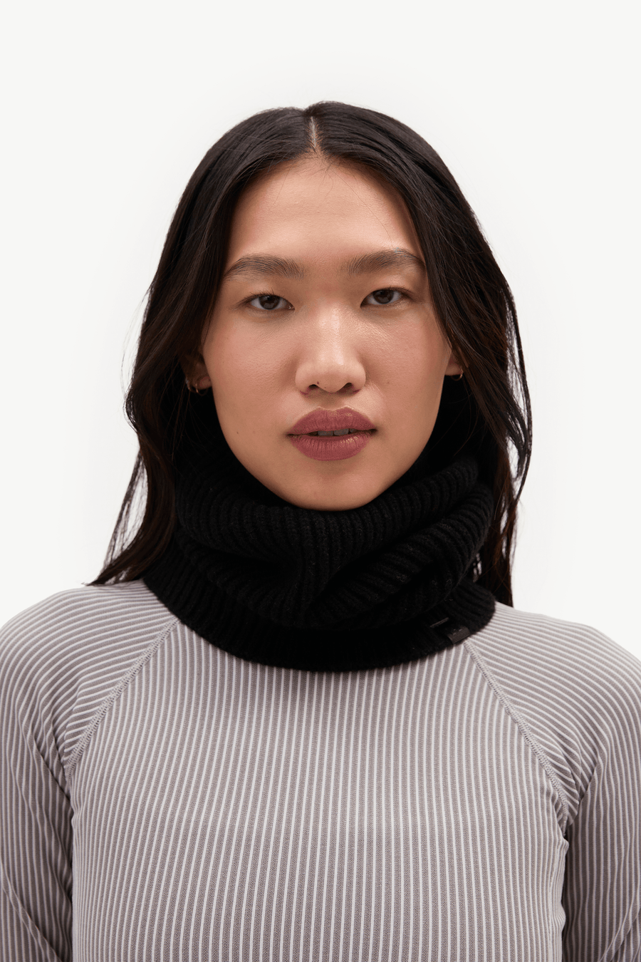 A model wearing a black ribbed knit cowl neck warmer over a gray and white vertically striped long-sleeve top, photographed against a clean white background.