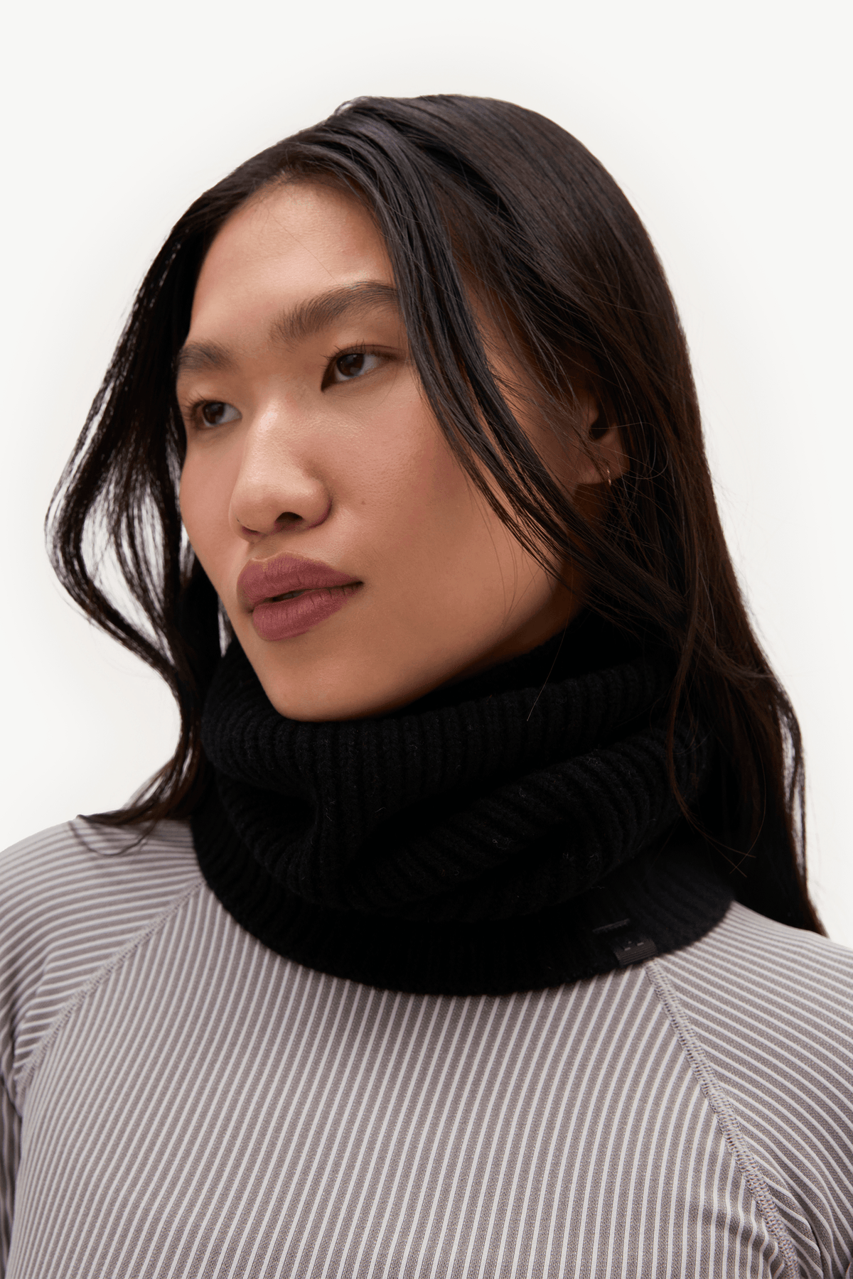 A close-up portrait of a woman wearing a thick, ribbed black turtleneck sweater over a gray and white vertically striped top, set against a plain white background.