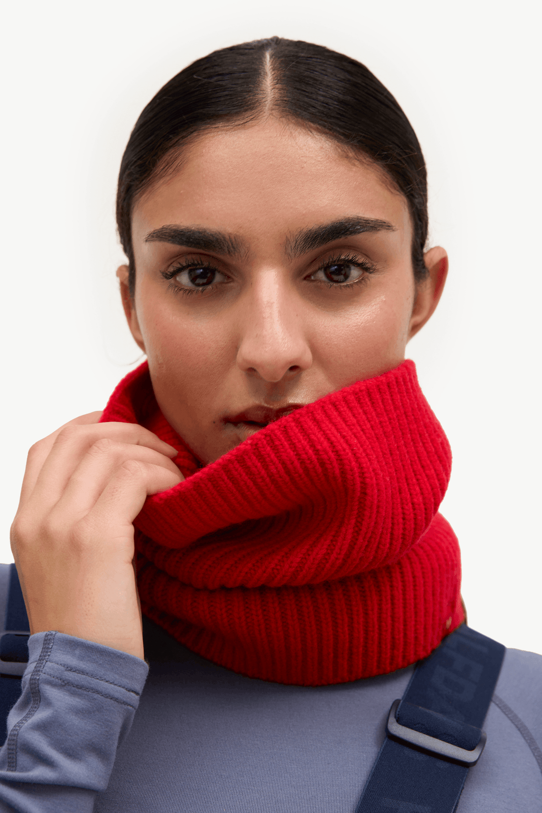 A close-up of a model wearing a bright red ribbed knit cowl neck warmer over a slate blue top, against a clean white background.