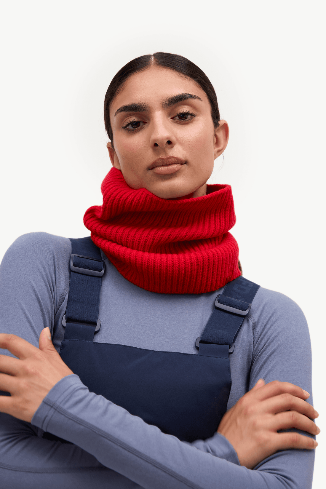 A woman wearing a bright red ribbed knit cowl neck warmer over a blue-gray long-sleeve top and navy overalls, posed against a clean white studio background.