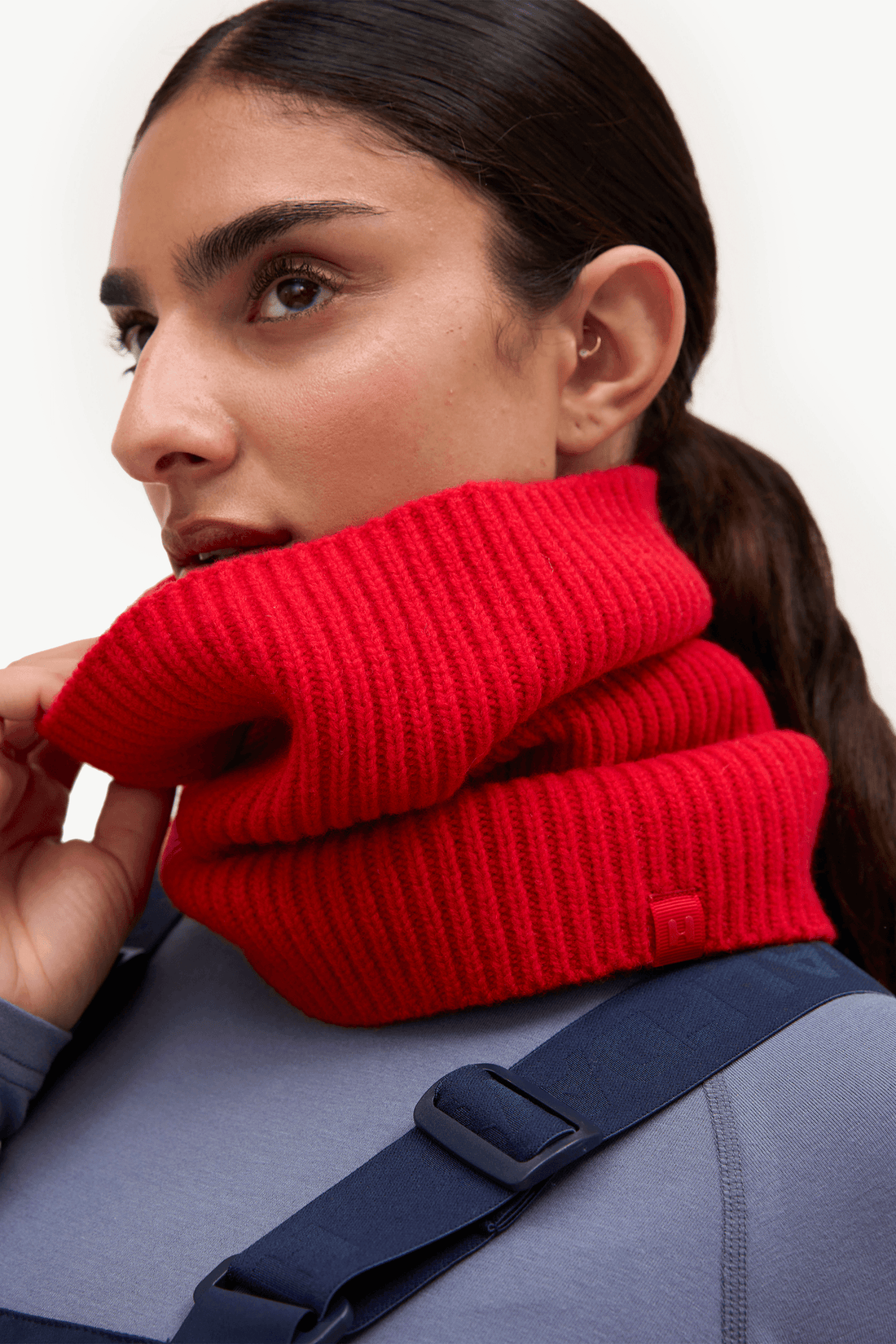 A close-up of a woman wearing a bright red ribbed knit cowl neck warmer, adjusting it around her neck. She is wearing a slate blue-gray top with a navy strap across the chest, set against a clean white background.