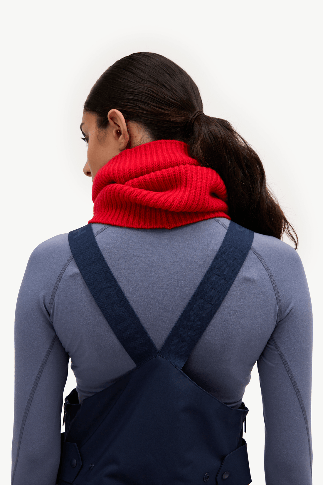 A woman shown from the back wearing a bright red ribbed knit neck warmer over a gray long-sleeve top and navy bib overalls, against a plain white studio background.