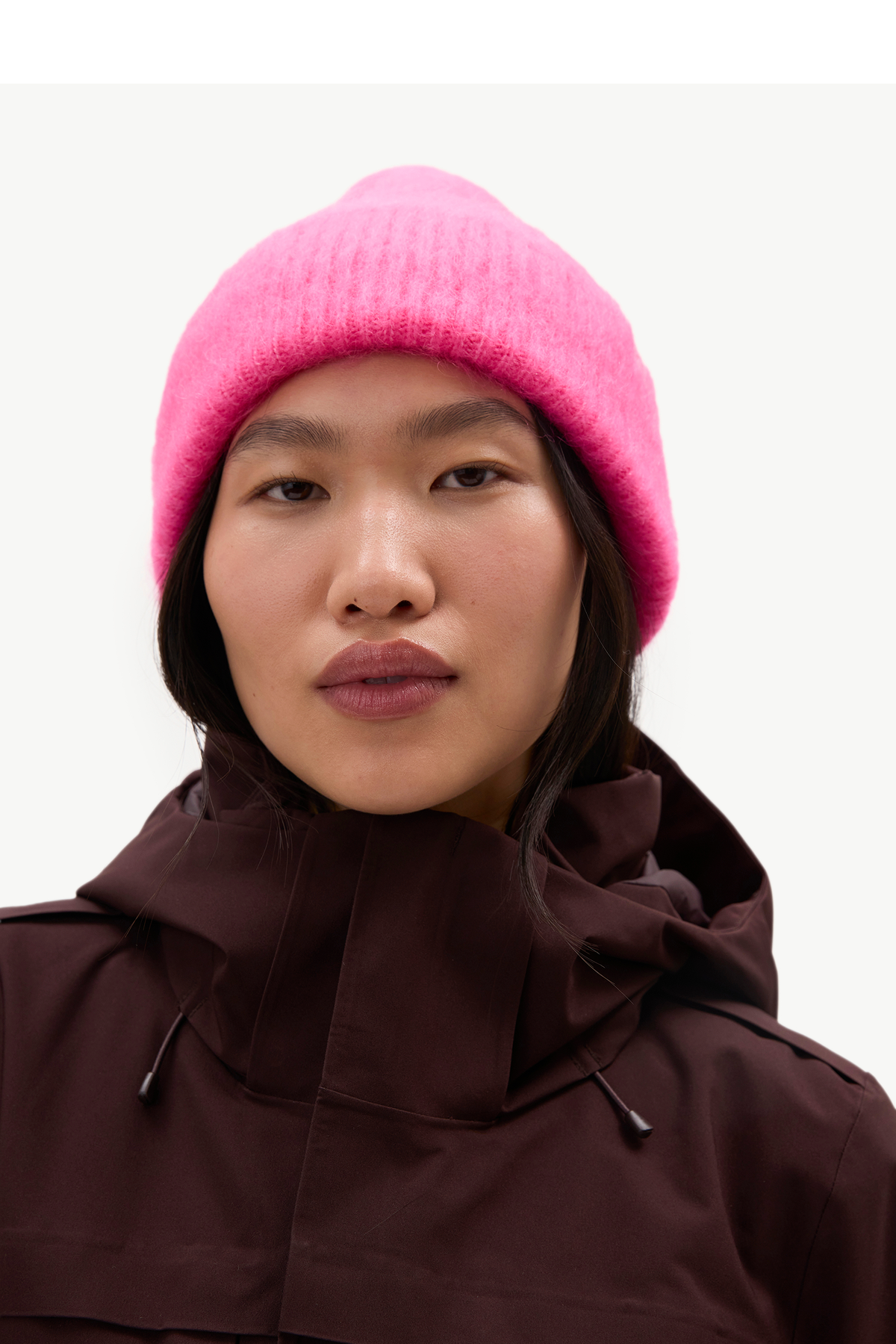 A close-up of a model wearing a bright pink fuzzy knit beanie and a dark brown hooded jacket against a plain light background.