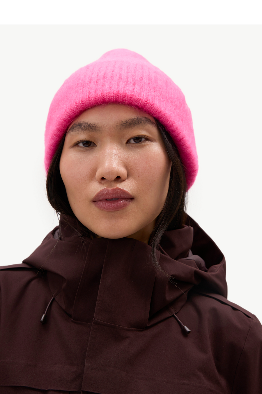 A close-up of a model wearing a bright pink fuzzy knit beanie and a dark brown hooded jacket against a plain light background.
