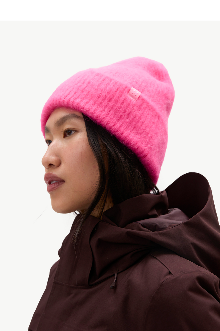 A close-up side profile of a person wearing a bright pink fuzzy knit beanie and a dark brown hooded jacket against a plain light background.