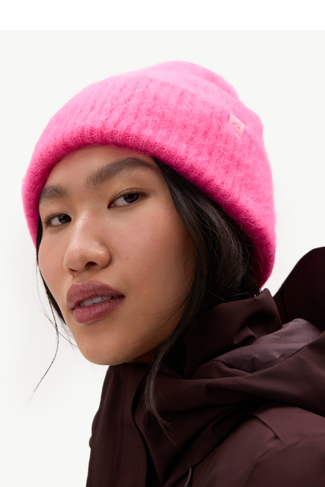 A close-up of a model wearing a bright pink fuzzy knit beanie and a dark brown hooded jacket against a plain light background.