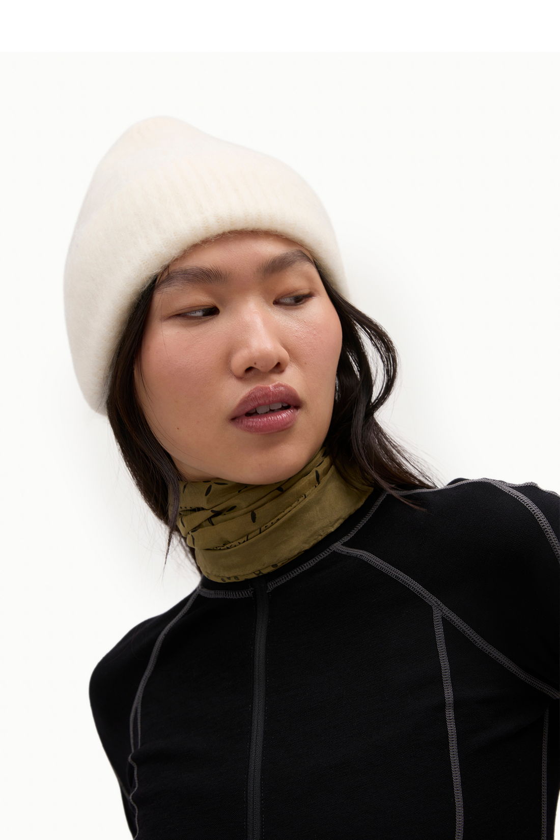 A model wearing a cream ribbed knit beanie, an olive green patterned neck gaiter, and a black athletic top against a plain white background.