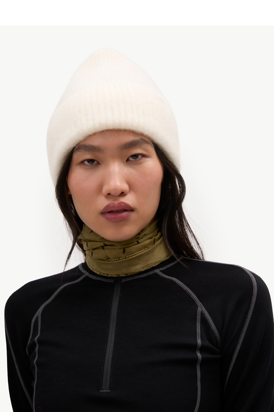 A model wearing a white ribbed knit beanie, an olive green neck gaiter, and a black long-sleeve quarter-zip athletic top with gray piping, set against a plain white studio background.