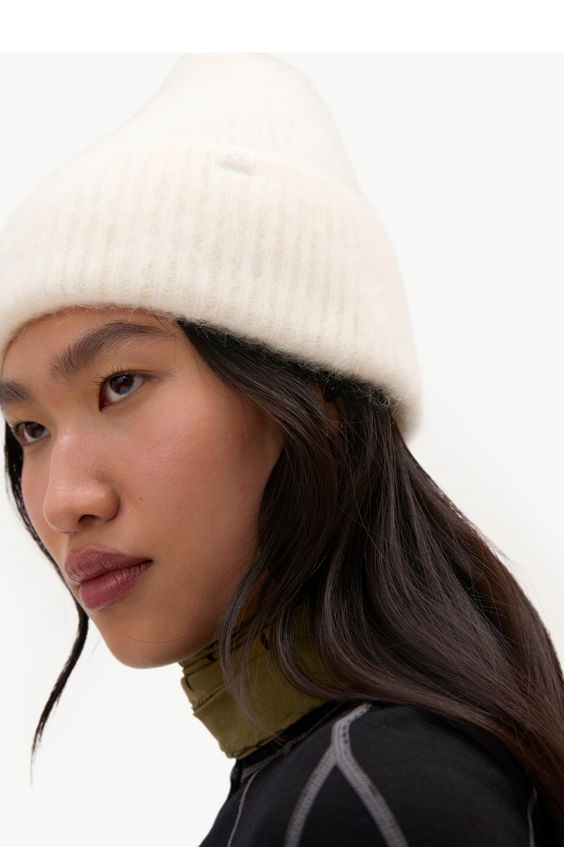 A close-up of a model wearing a cream-colored ribbed knit beanie hat, paired with a dark olive green neck warmer and a black top against a plain white background.