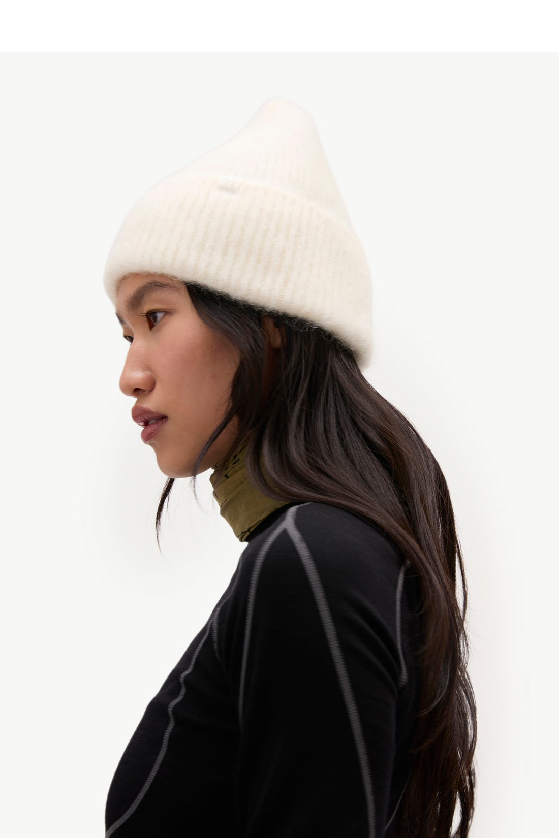 A woman in profile wearing a cream ribbed knit beanie, a black athletic top, and an olive green neck gaiter, set against a plain light gray studio background.