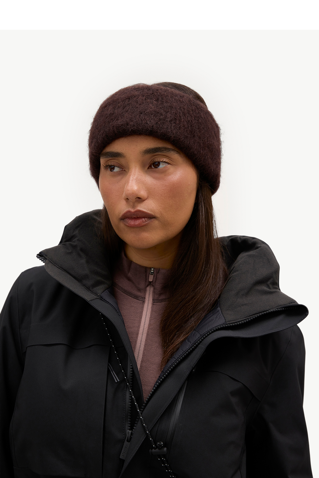 A woman wearing a dark brown fuzzy knit beanie and a black hooded jacket over a mauve zip-up top, photographed against a plain light background.