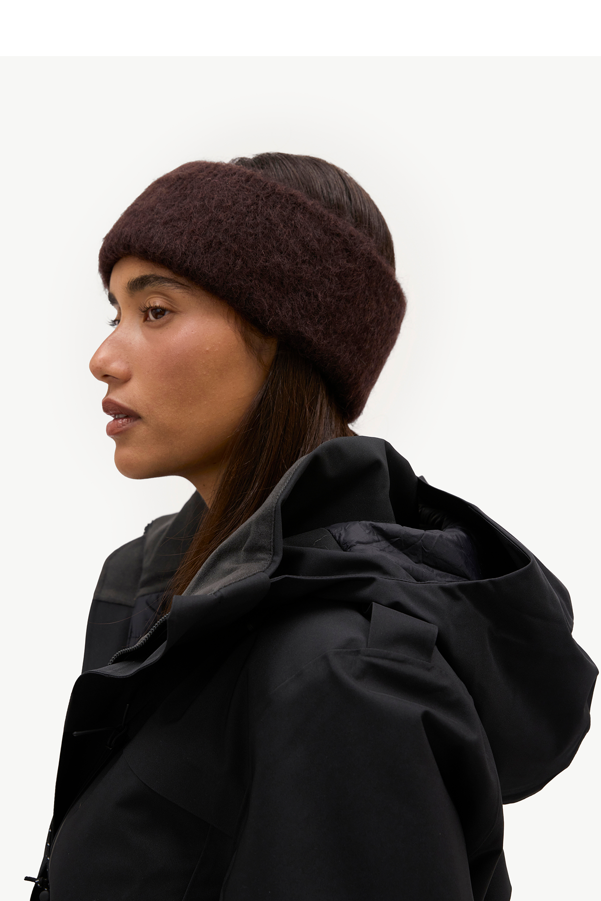 A woman in profile wearing a dark brown fuzzy knit beanie and a black hooded jacket against a plain light background.