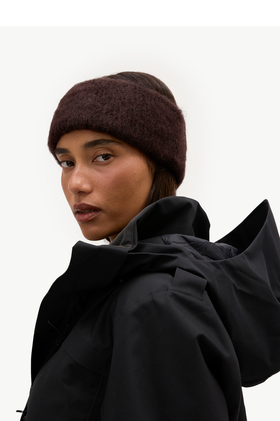 A close-up of a model wearing a dark brown fuzzy knit beanie and a black hooded jacket against a plain light background.