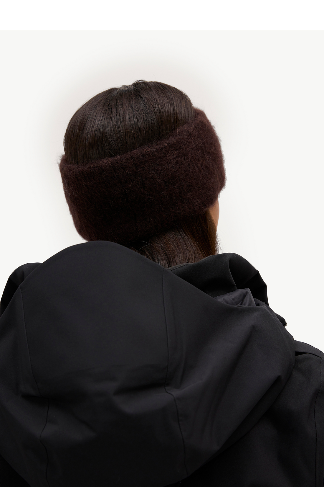 A person shown from behind wearing a thick, fuzzy dark brown ear warmer headband and a black hooded jacket against a plain white background.