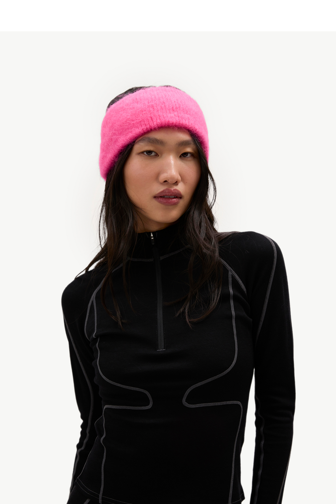A woman wearing a bright pink fuzzy knit headband and a black long-sleeve quarter-zip athletic top with gray contrast stitching, photographed against a plain light background.