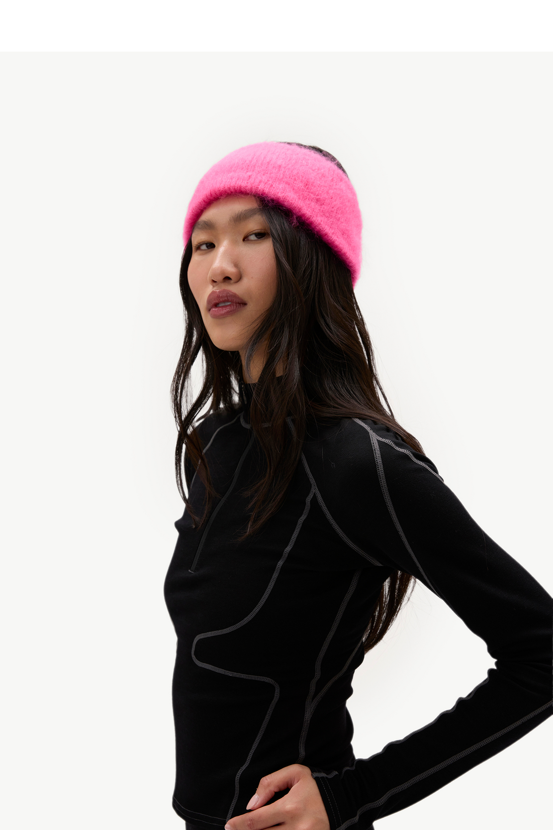 A woman wearing a bright pink fuzzy knit headband and a black athletic long-sleeve top with gray seam details, photographed against a plain light background.