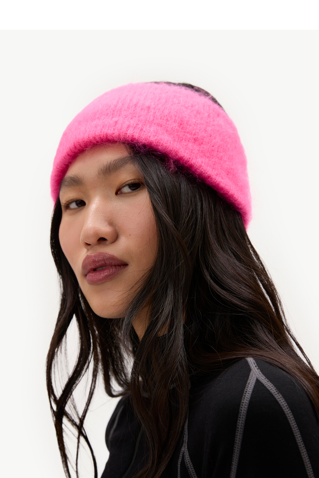A close-up of a model wearing a bright pink fuzzy knit headband against a plain white background.