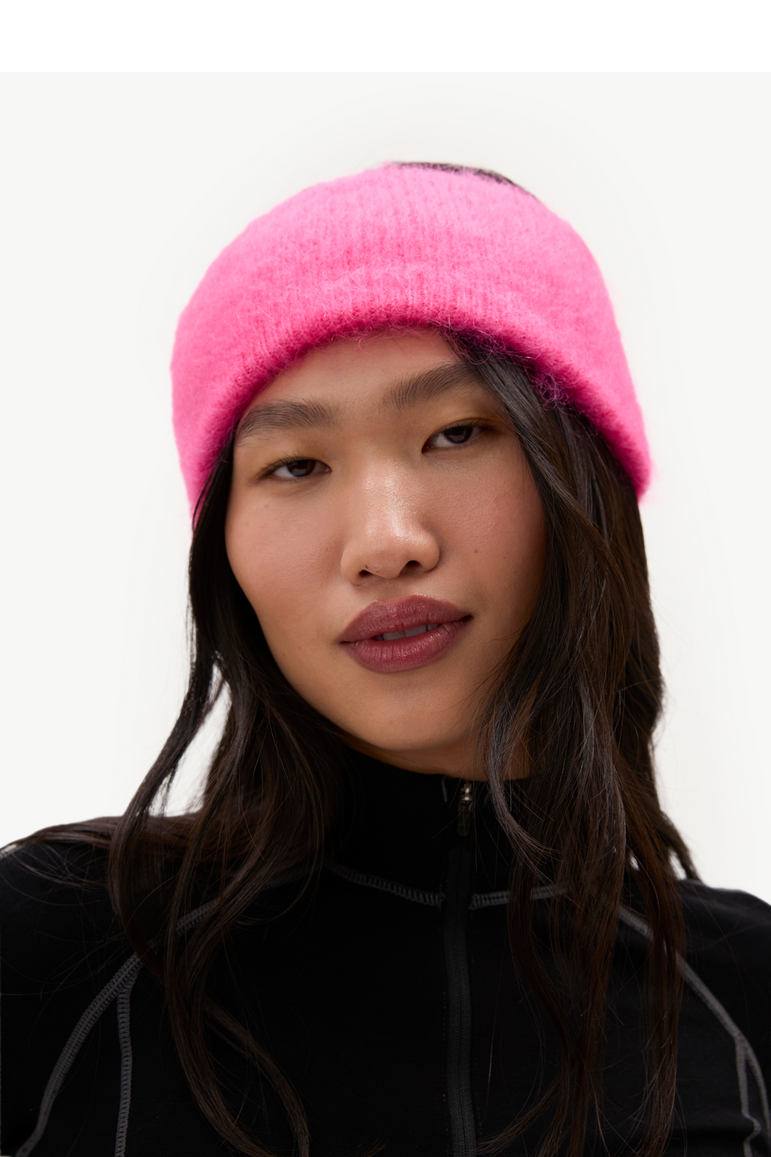 A close-up of a model wearing a bright pink fuzzy knit beanie and a black zip-up top against a plain light background.