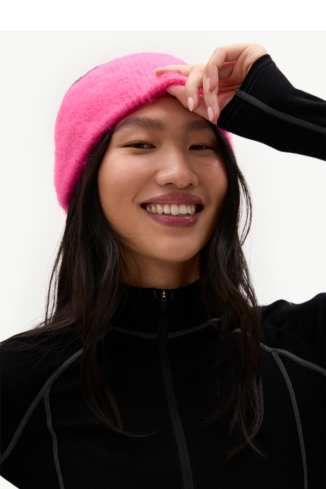 A smiling woman wearing a bright pink fuzzy beanie and a black zip-up athletic jacket, photographed against a clean light gray studio background.