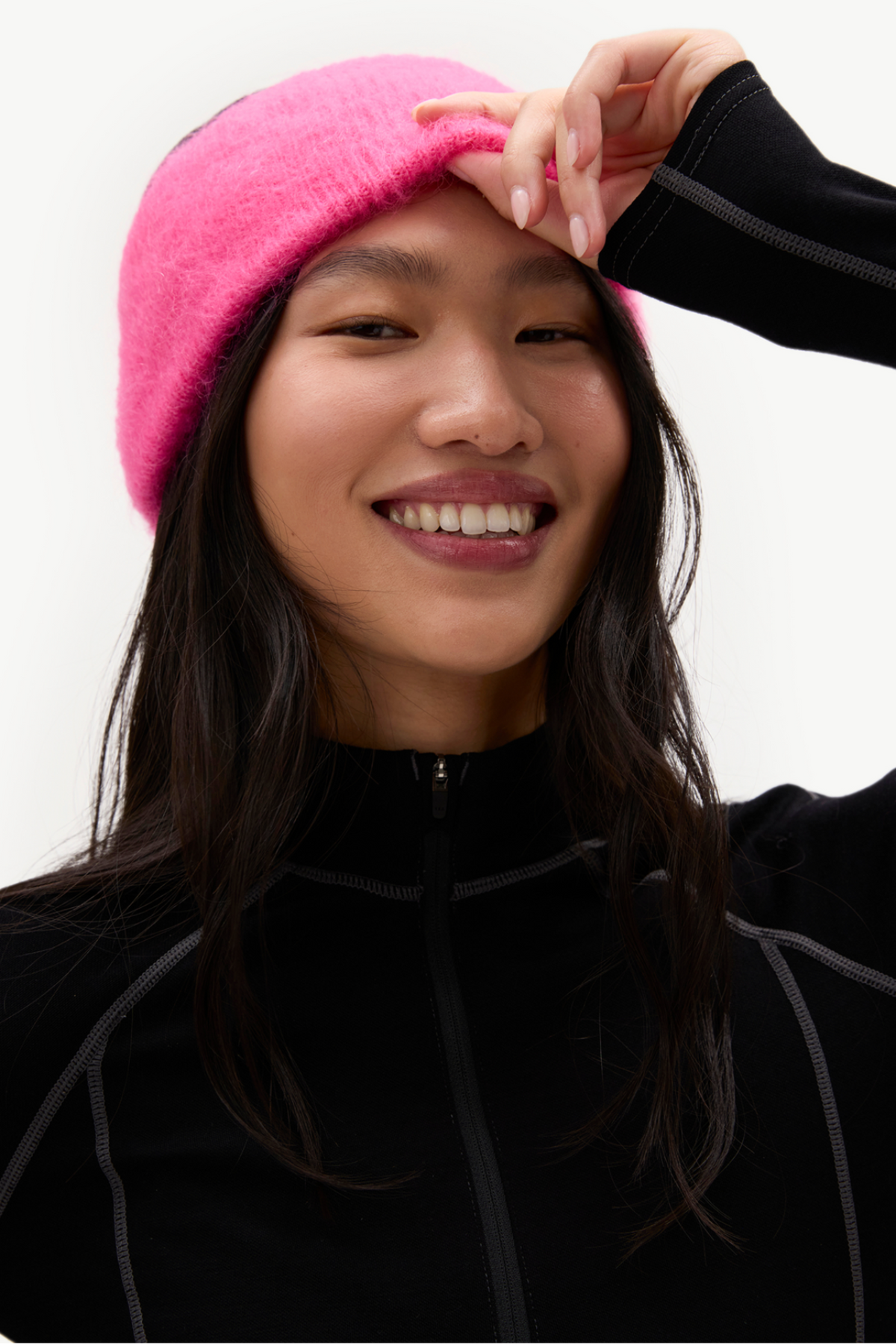A smiling woman wearing a bright pink fuzzy beanie and a black zip-up athletic jacket, photographed against a clean light gray studio background.