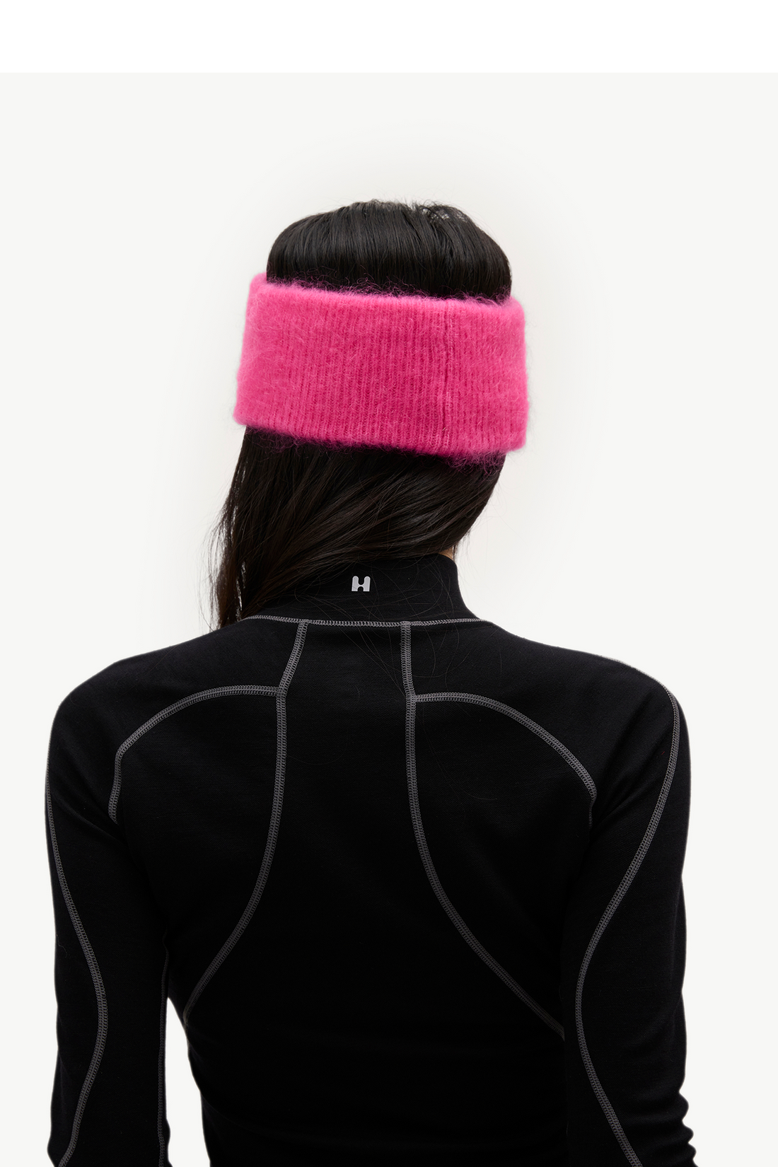 A person shown from the back wearing a bright pink fuzzy knit headband and a black athletic long-sleeve top with gray seam detailing, against a plain light background.